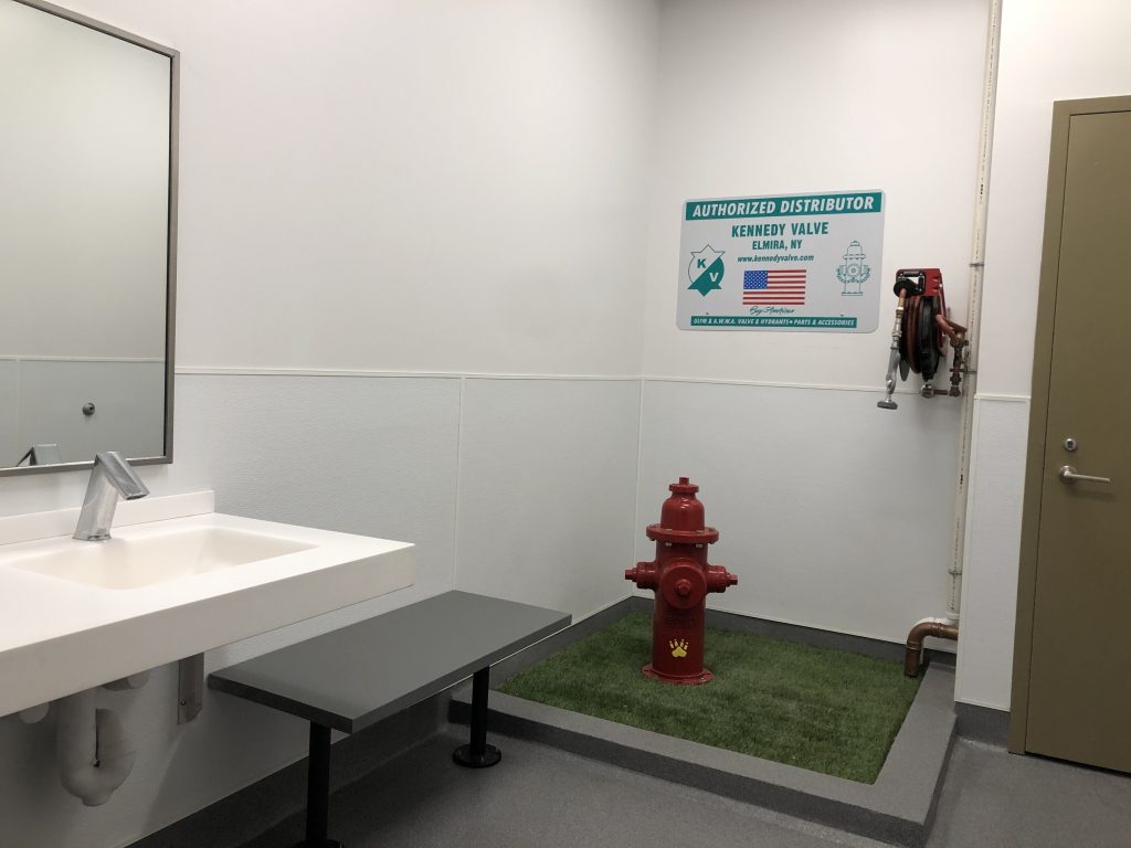 Animal relief area inside an airport consists of a red fire hydrant placed on a patch of artificial turf with a nearby coiled hose, bench, sink, and trash and compost.