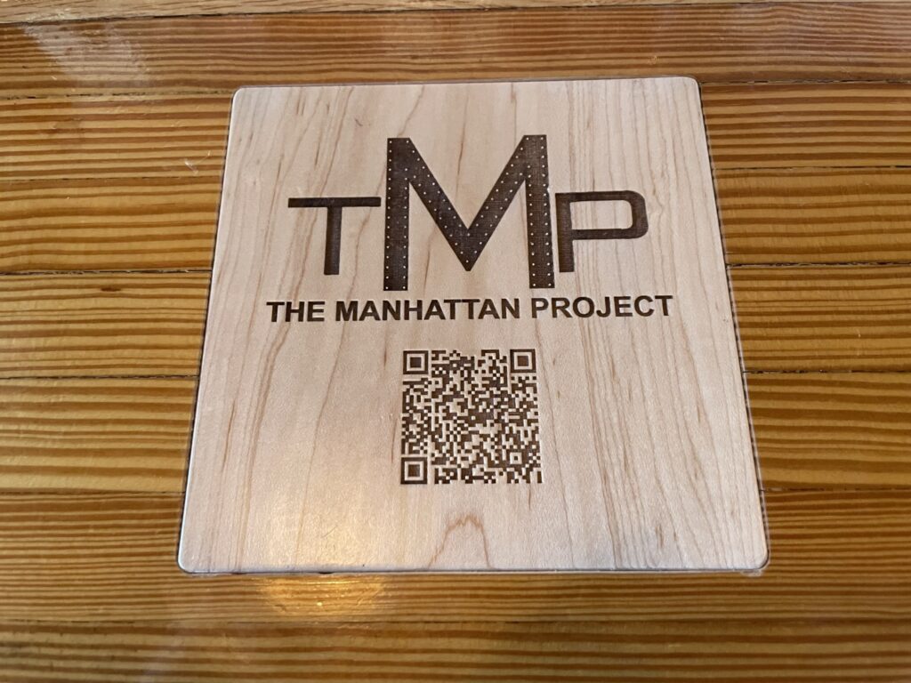 A light coloured wooden square is inset into a darker coloured wooden barton. The inset contains the letters TMP, followed by the words "The Manhattan Project", followed by a QR code. All 3 pieces of content are etched or laser cut into the surface of the wood and the deep groves are darker in colour providing a high contrast distinction against the un-etched surface. The etching is at a depth that facilitates tactile consumption of the content.