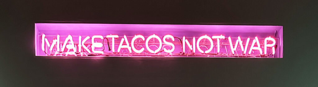 A florescent pink neon sign set against a dark background reads "Make tacos not war